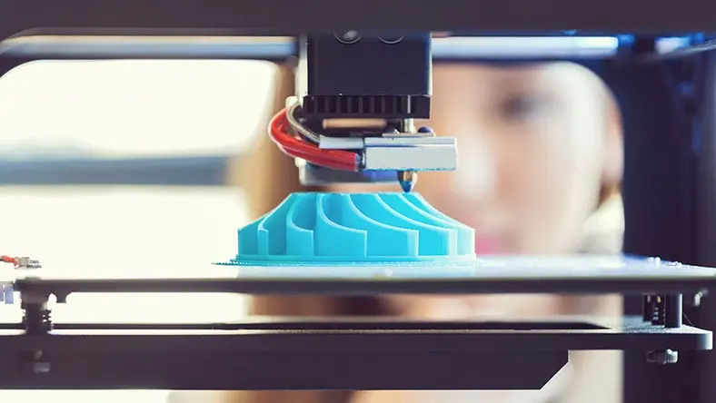 A young woman watches a printout on a 3D printer.