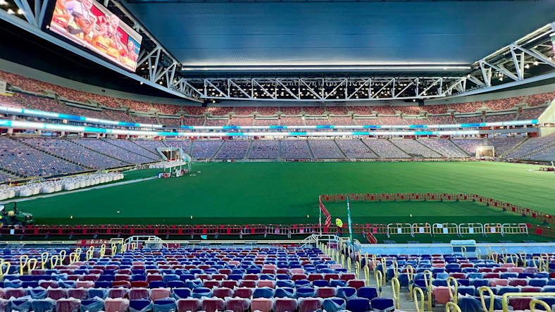 An empty stadium has covered seats and a large screen displaying a live video feed.