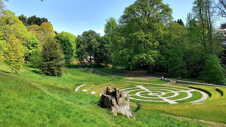 A park contains a circular stone labyrinth, an amphitheater, and many trees.