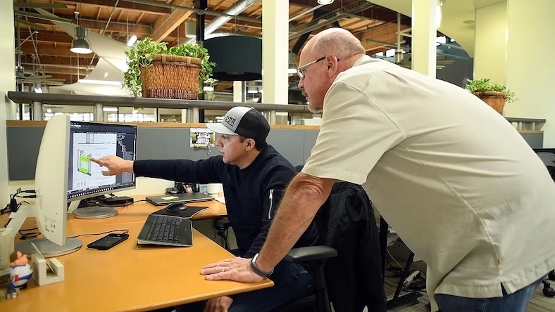 Two men in a loft office look at a computer monitor.