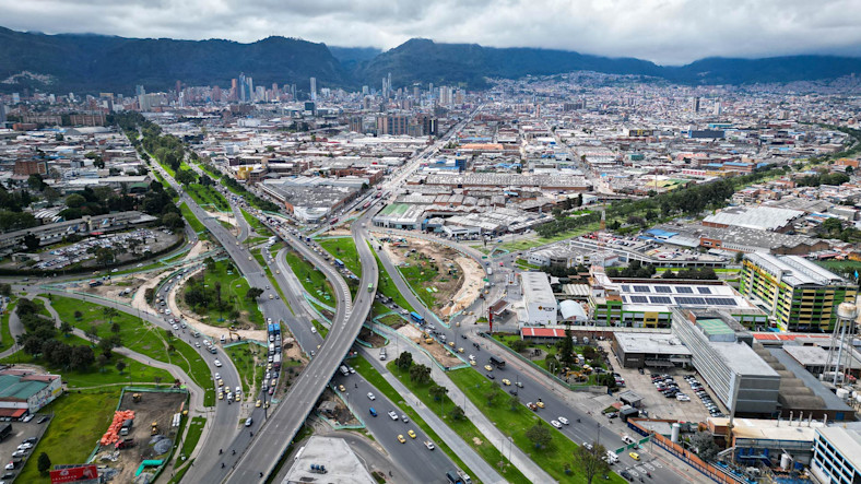 An aerial view shows a busy highway interchange in an urban cityscape with surrounding industrial and commercial buildings, green spaces, and distant mountains.