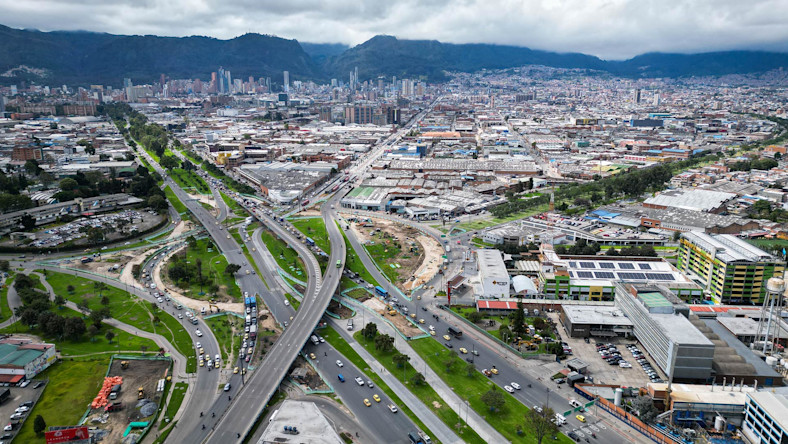 Conconcreto forges a digital path to sustainable infrastructure in Colombia An aerial view shows a busy highway interchange in an urban cityscape with surrounding industrial and commercial buildings, green spaces, and distant mountains.