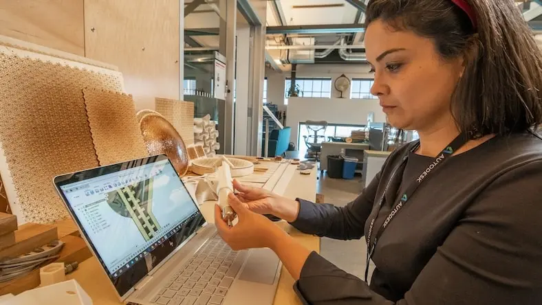 A woman holds a small manufactured piece while looking at a computer showing a 3D design in Fusion. 