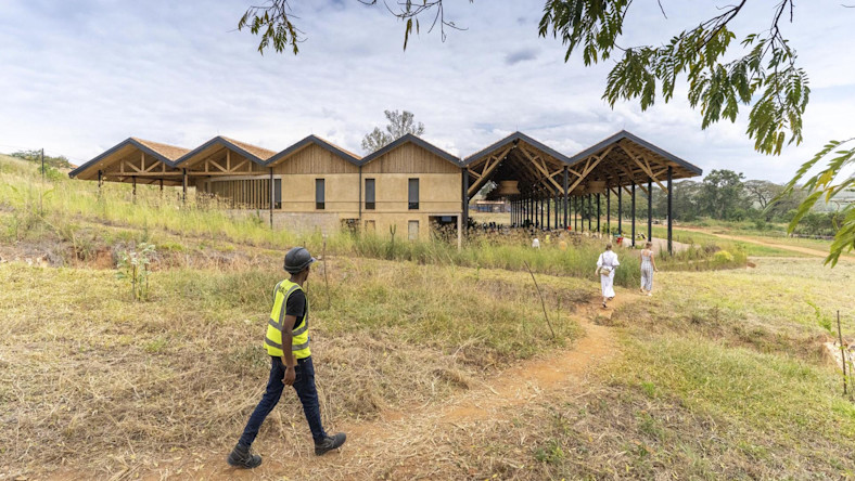 A worker in safety gear walks toward a wooden structure under construction in a rural area.