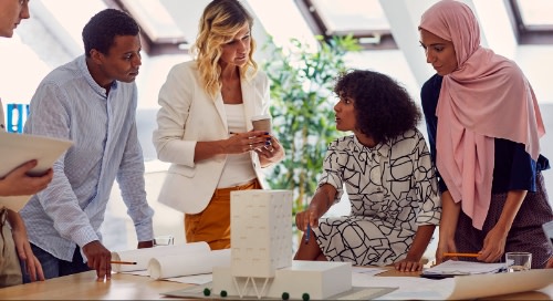 A multicultural group of architects stands around plans on a conference table.