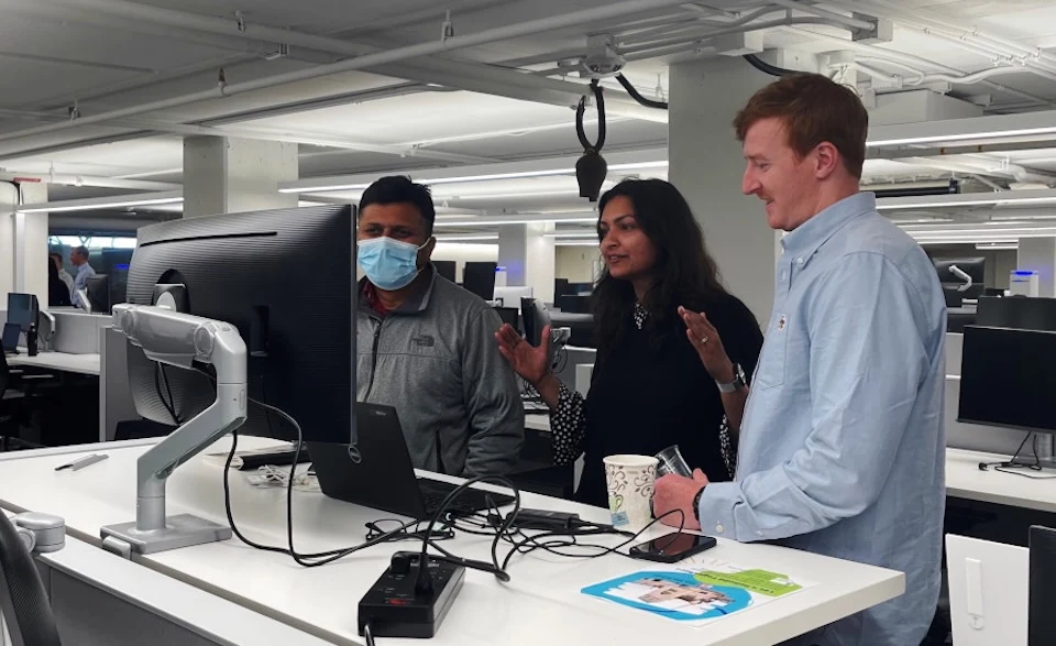 Sue Bhattacharjee and two male colleagues look at a computer monitor at an office workstation.
