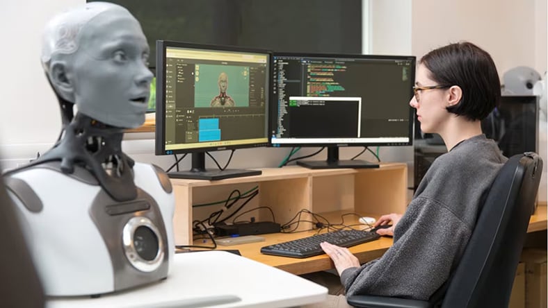 a woman sits at a desk with a robot head model and a computer.