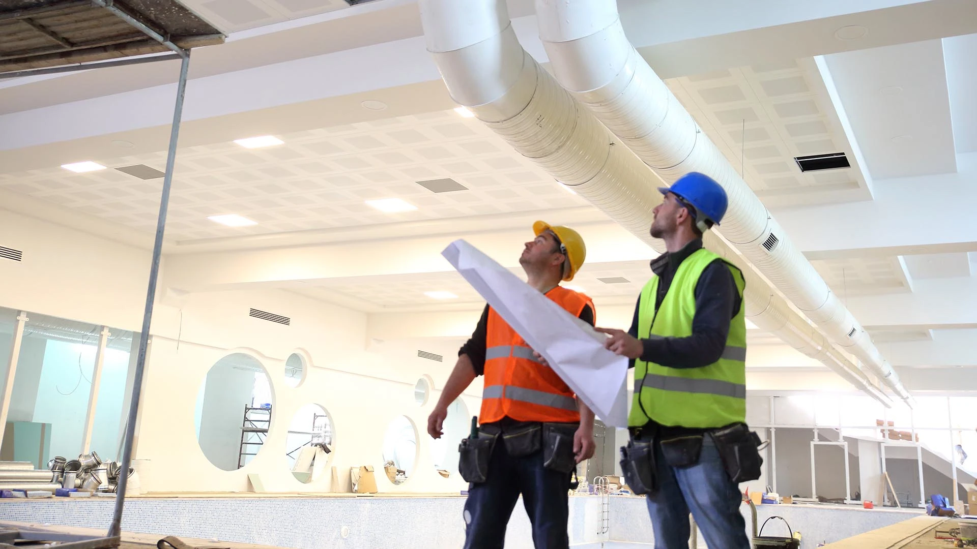 Dos operarios examinan el sistema de climatización en una obra de interior, con planos en la mano y la mirada en alto.