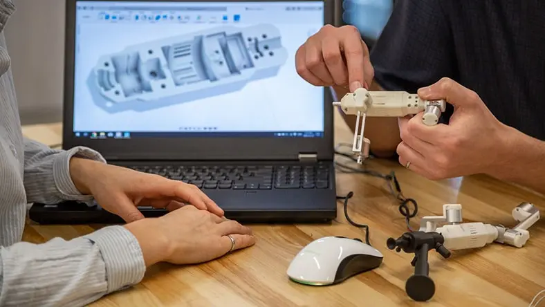 Two people assembling a 3D-printed mechanical component while referencing a CAD design displayed on a laptop screen.