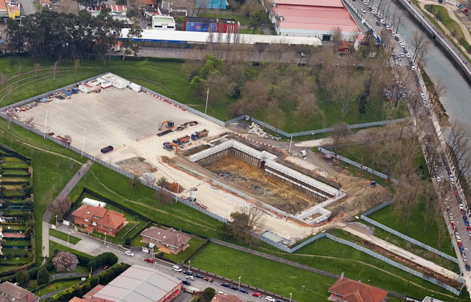 Aerial view of the Hermanos Castro storm tank under construction in Gijón, showing the excavation site within an urban park and surrounding drainage infrastructure.