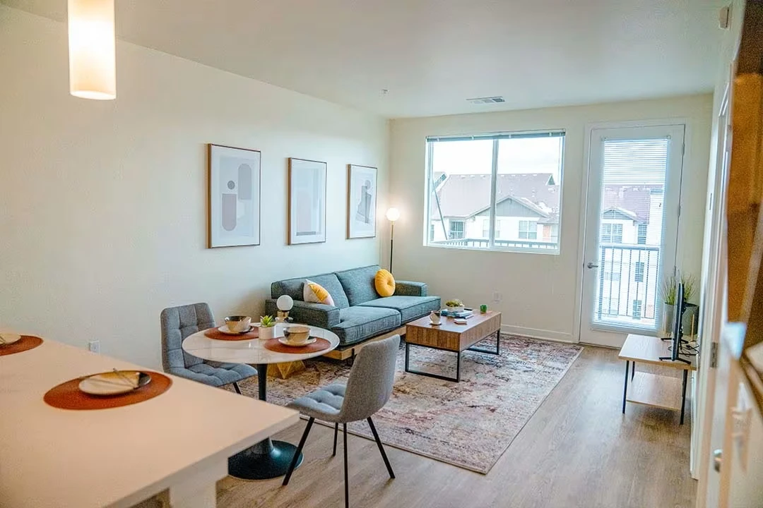 Bright, modern living room with a sofa, coffee table, dining nook, framed art, and sliding glass door to a balcony.