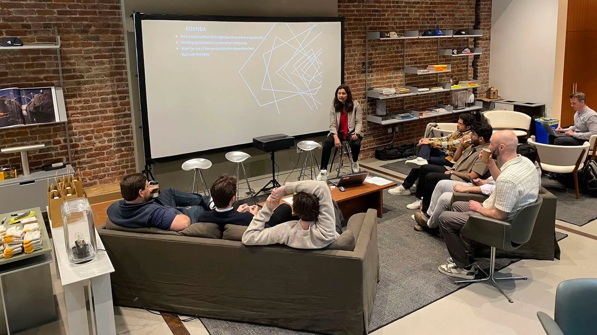Sue Bhattacharjee sits in front of a projector screen talking to several men in the common area in the Formwork Labs office.
