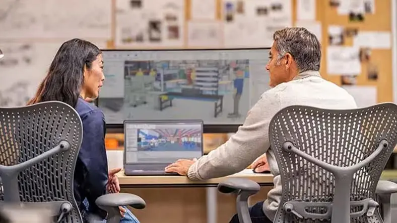 Two coworkers sit in office, looking at computer screen.