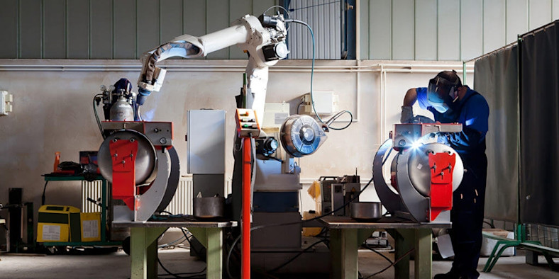 A welder in a factory works next to a large industrial robot.
