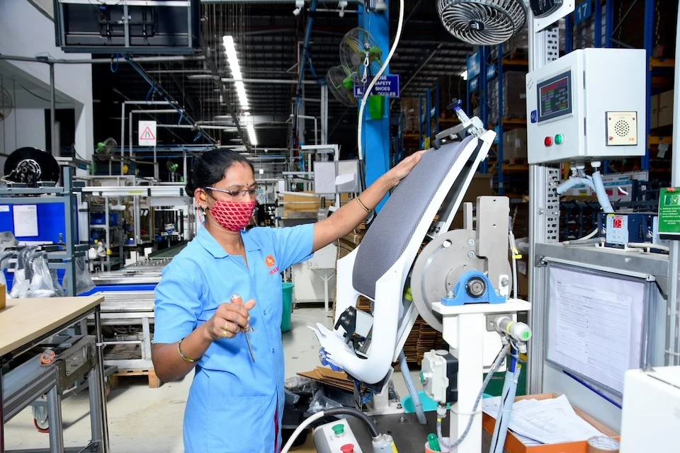 In India, a woman in a furniture factory operates a machine that measures a chair seat.