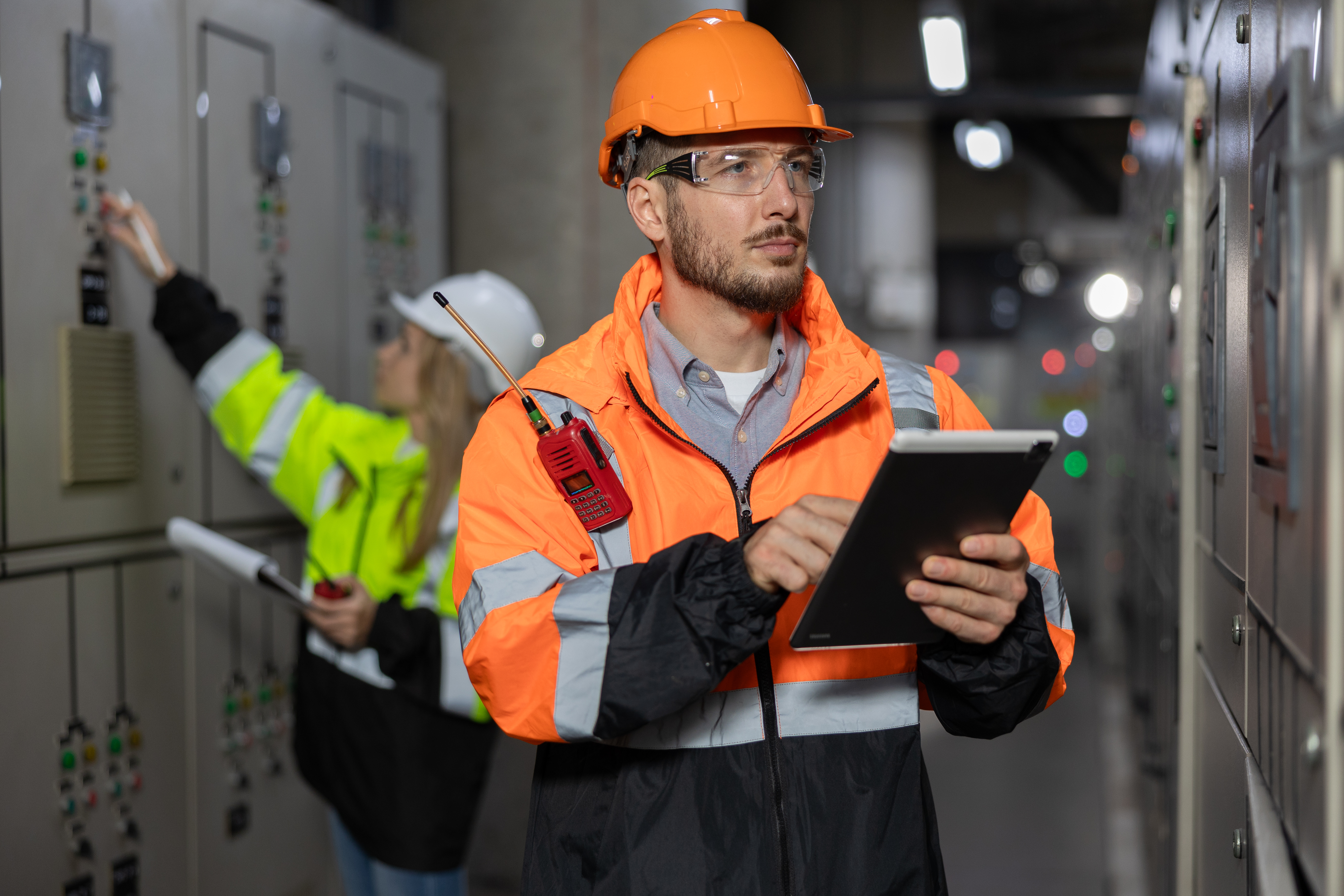 A male worker in a safety jacket is holding a tablet and inspecting control panels, while a female worker stands in the background.  