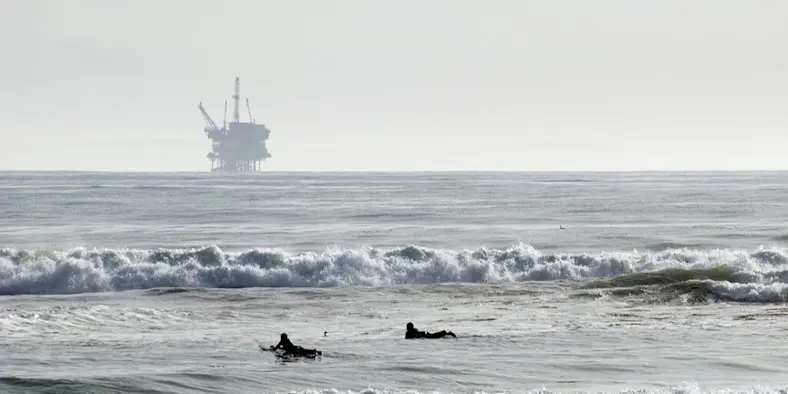Surfers paddle out into the ocean, with an oil rig in the distance
