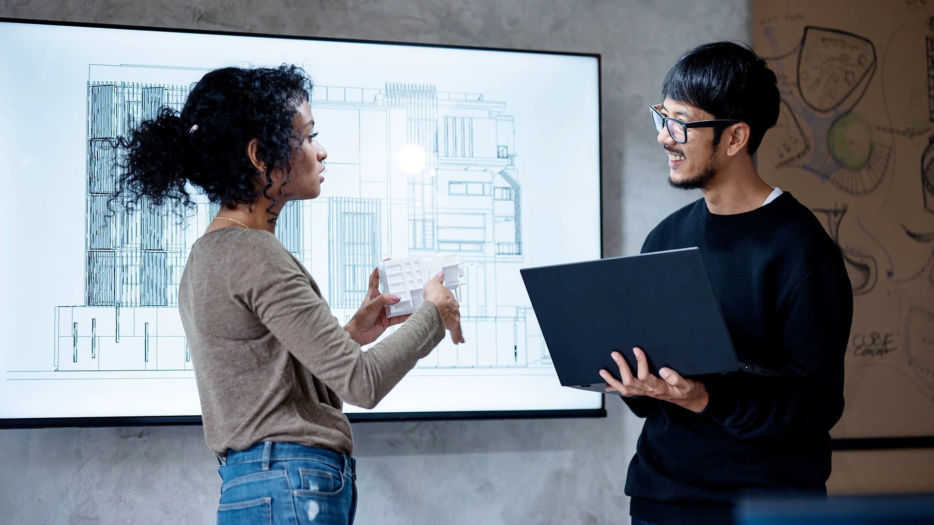 Two workers discuss a building model design on a large screen on the wall behind them. 