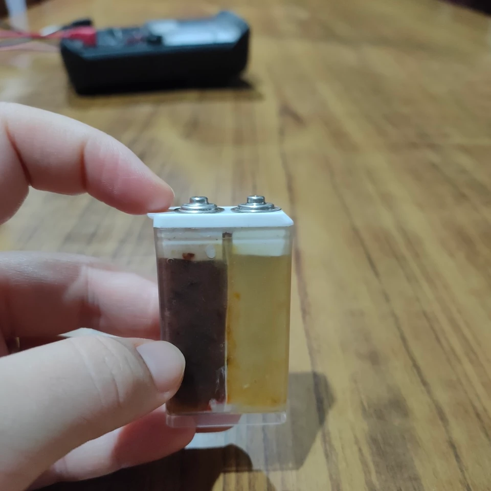 A hand holds a small battery cell prototype with materials visible through the transparent case.