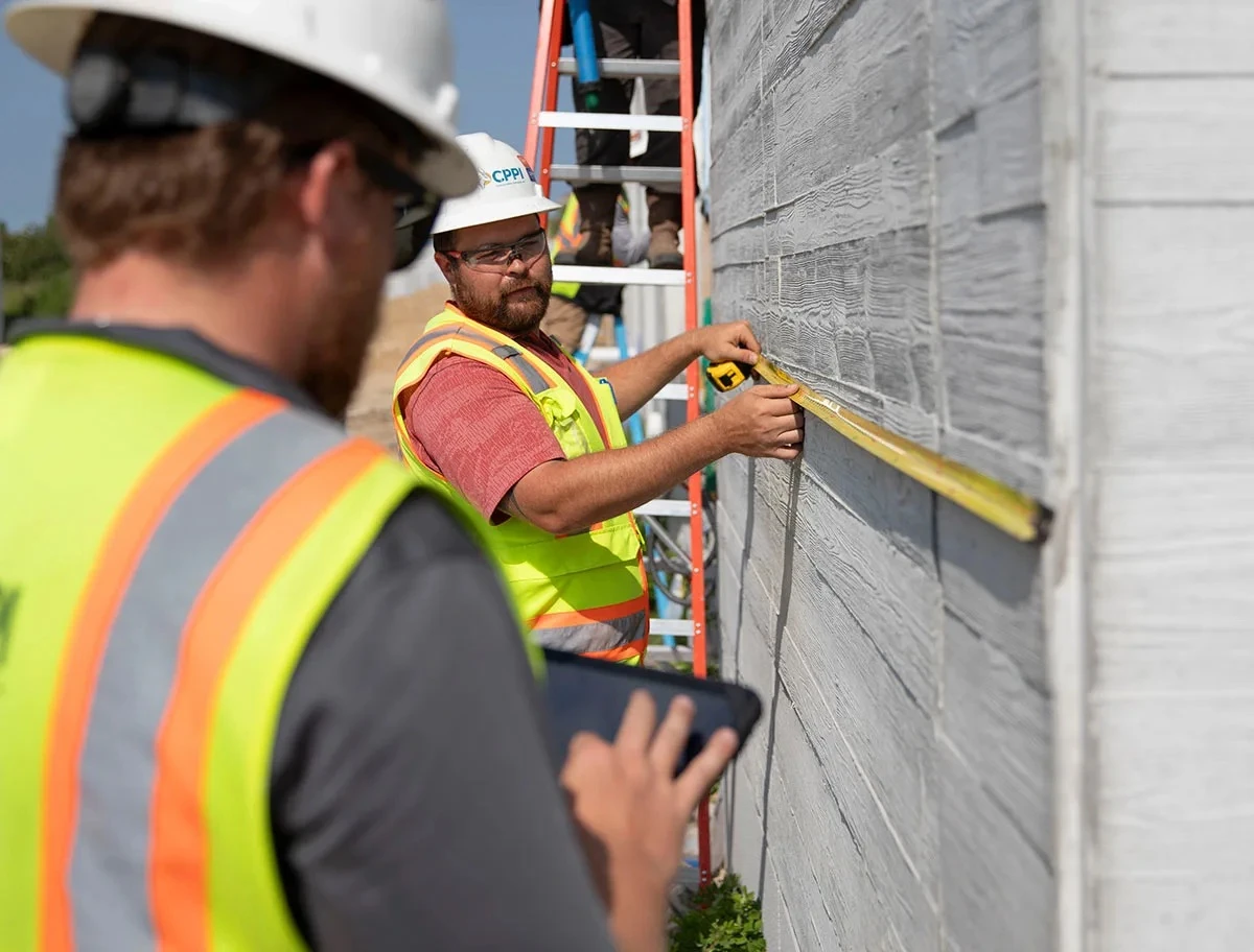Construction worker measures an exterior wall with a tape while a colleague records details on a tablet.