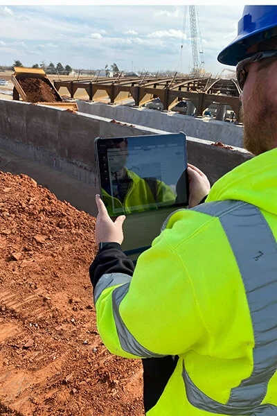 A man in a safety jacket holds a tablet on a jobsite