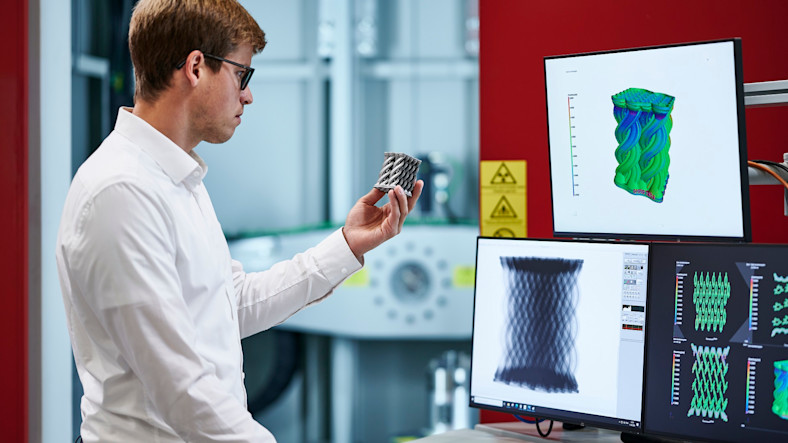 A man holds a small, textured object and compares it with digital images from scans and simulations displayed on screens