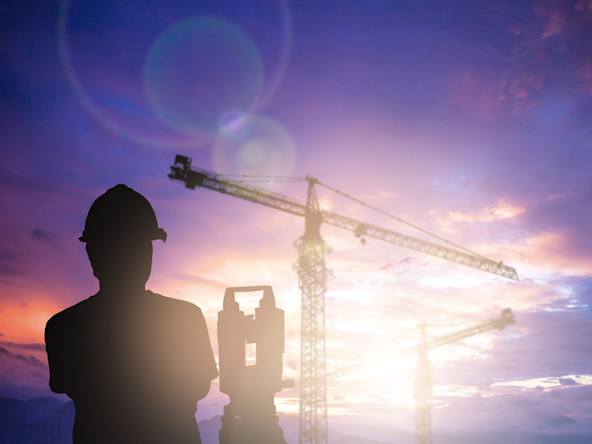 Silhouette eines Vermessungstechnikers vor einem unscharf abgelichteten Baukran bei Sonnenaufgang.