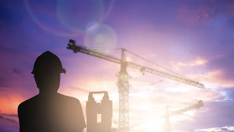 A silhouette of survey engineer working in a building site is over a blurred construction site at sunrise.
