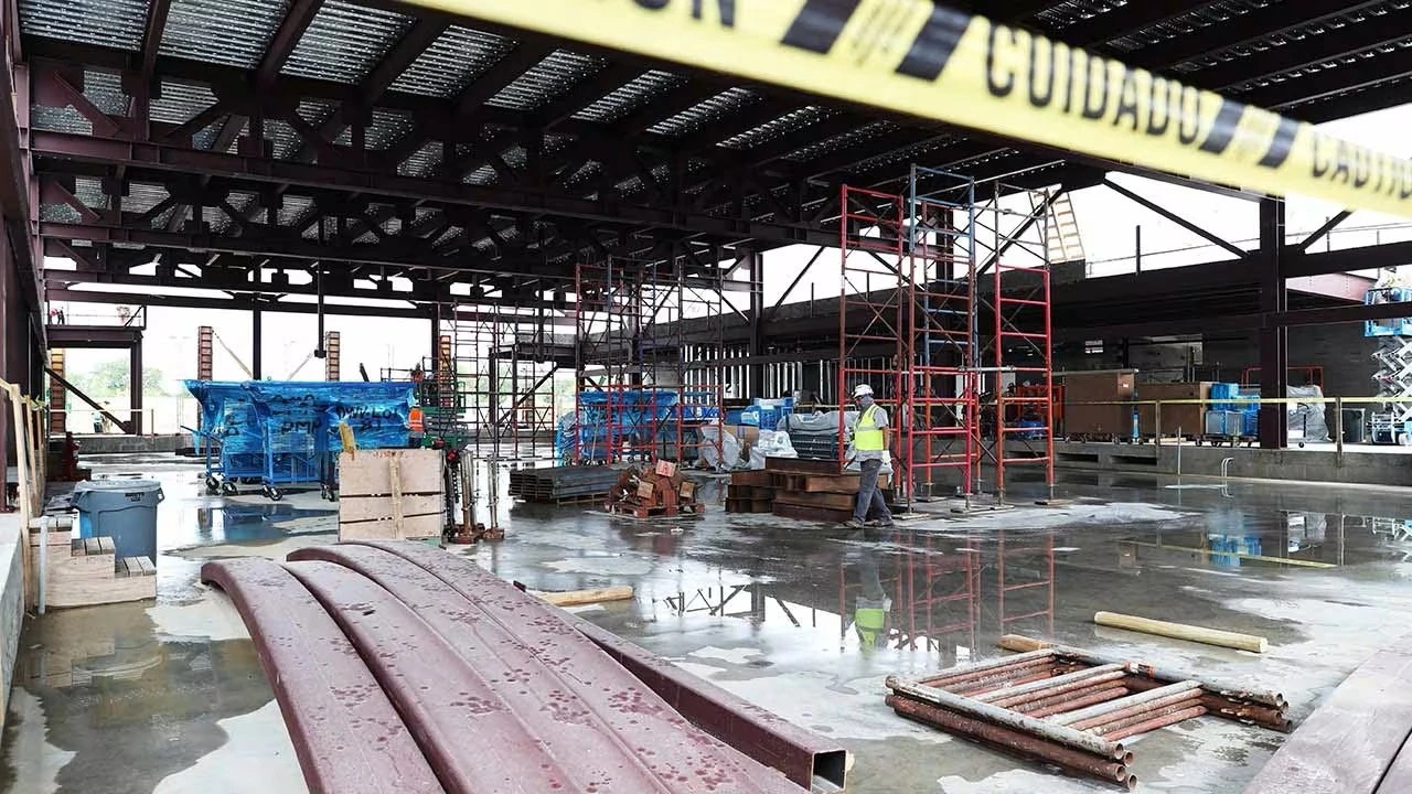 Interior of a large building under construction with steel beams, scaffolding, materials, and a worker walking across a wet concrete floor.