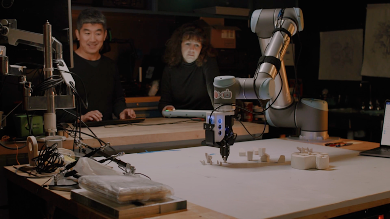 Two people watching a robotic arm 3d print pieces of coral for a coral reef