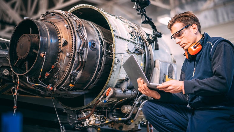 Aircraft engineer in a hangar using a laptop while repairing and maintaining an airplane jet engine