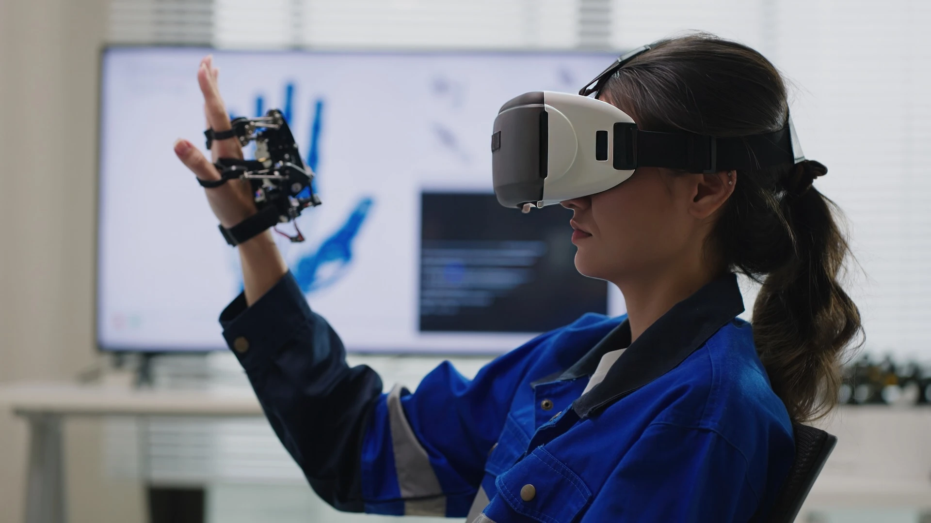 A woman wearing a VR headset uses a device strapped to her hand to interact with a robotics design program.