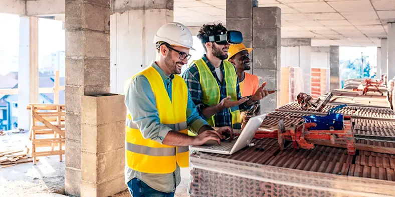 three construction workers on a jobsite using a laptop and VR goggles