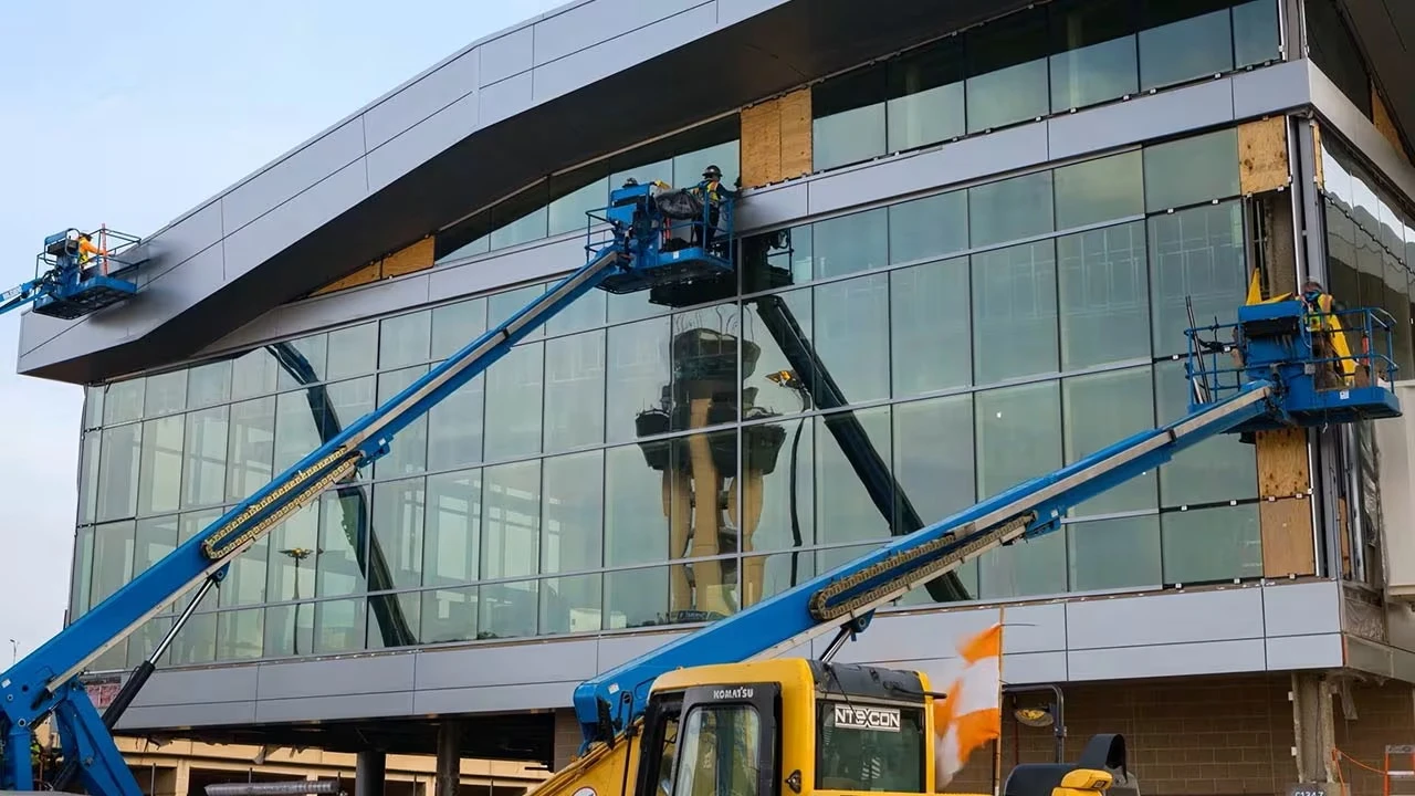 Workers use boom lifts to install large glass panels on a modern building facade under construction.