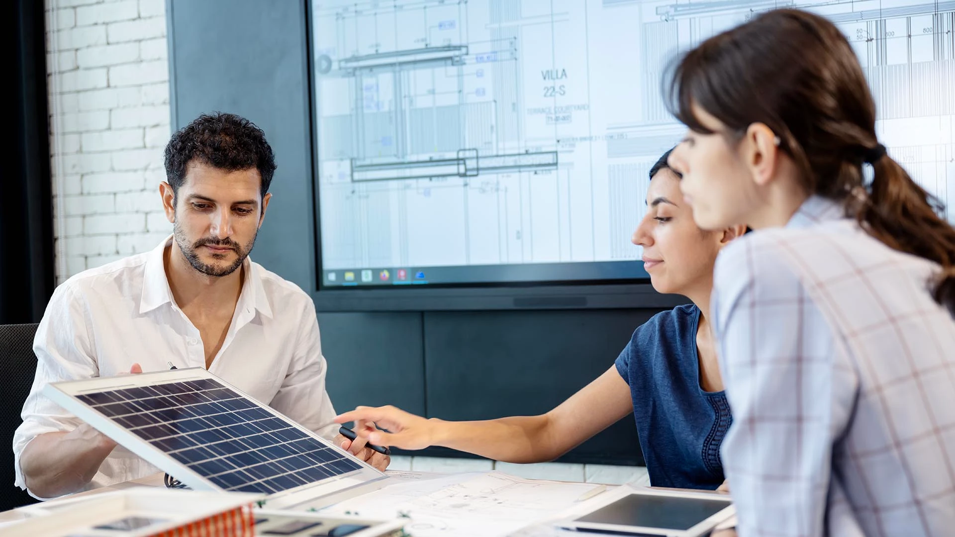 En una oficina moderna, tres expertos examinan un prototipo de panel solar mientras observan planos arquitectónicos en una gran pantalla.
