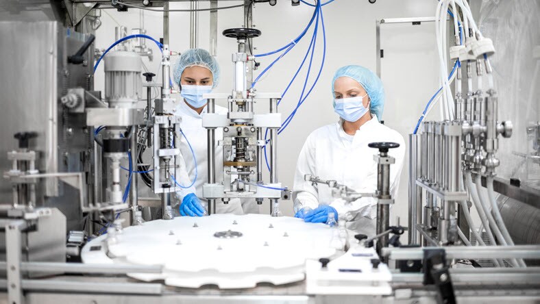 Two young female workers in protective gear operate a high-tech pharmaceutical machine that performs multiple functions including blowing, filling, capping, and labeling of bottles.