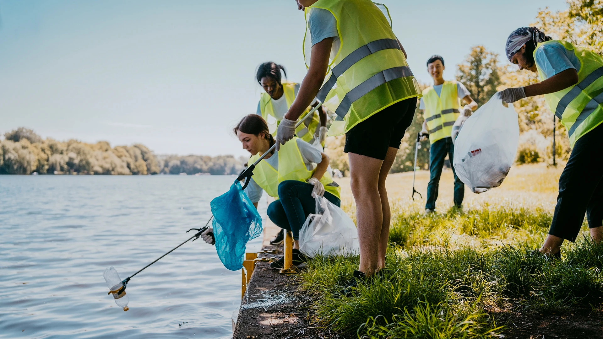 Several Gen Z environmentalist volunteers using grabber claws to pick up litter from park with lake