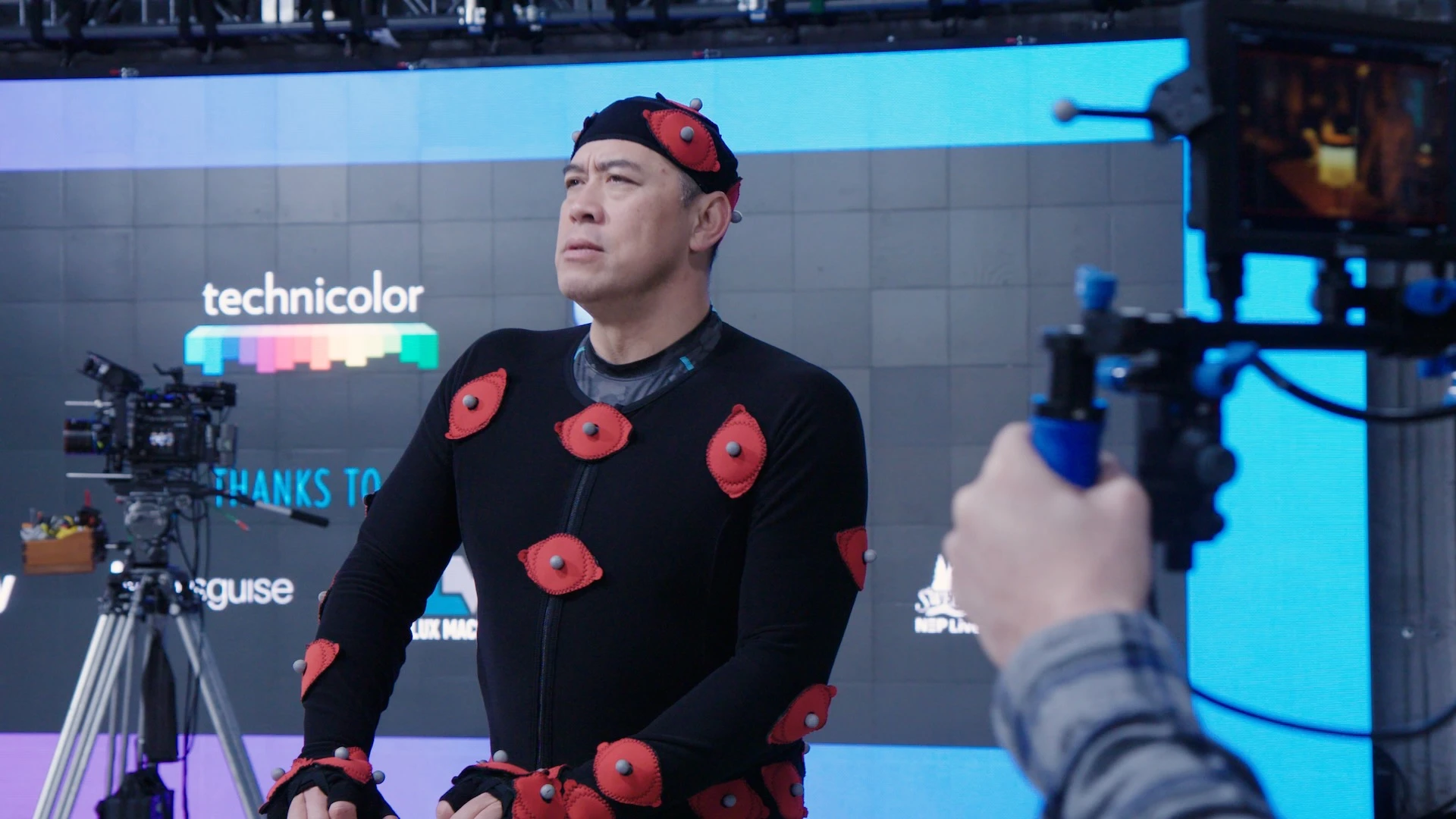 A man dressed in a sensor-covered motion capture suit and cap is being filmed by a camera crew.