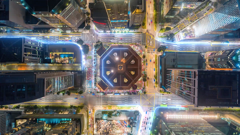 A top-down view of a brightly lit city intersection shows a large octagonal building at the center, surrounded by tall skyscrapers.