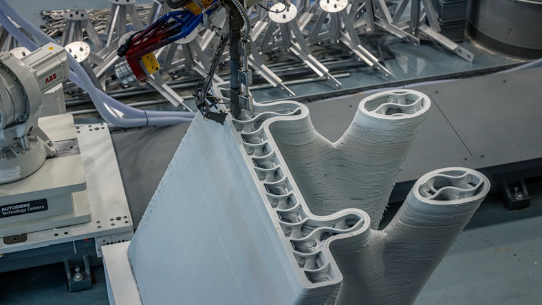 A robotic arm extrudes plastic material for a 3D-printed bridge test.