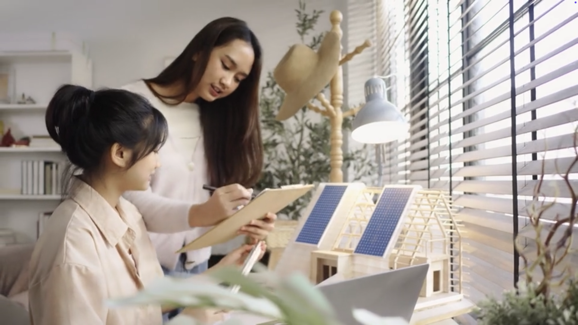 Two women discussing a project at a desk where there is a model of a house with solar panels on the roof.