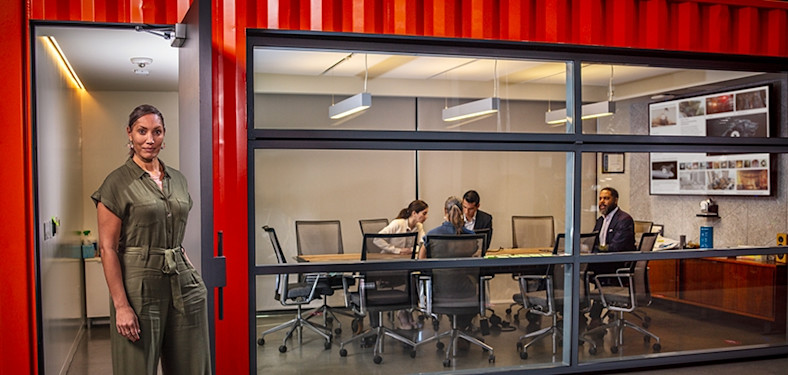 A woman stands in the doorway of a conference room where people are gathered around a table.