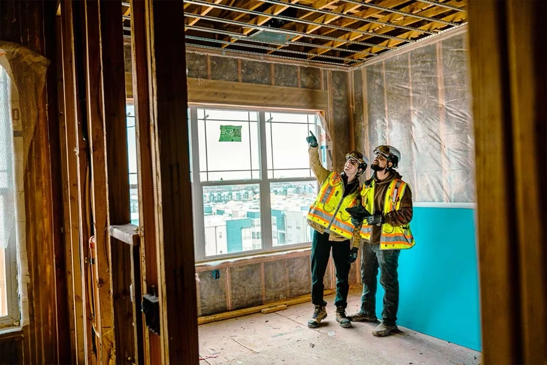 Two construction workers in safety vests inspect ceiling framing inside a partially built apartment with a large window.