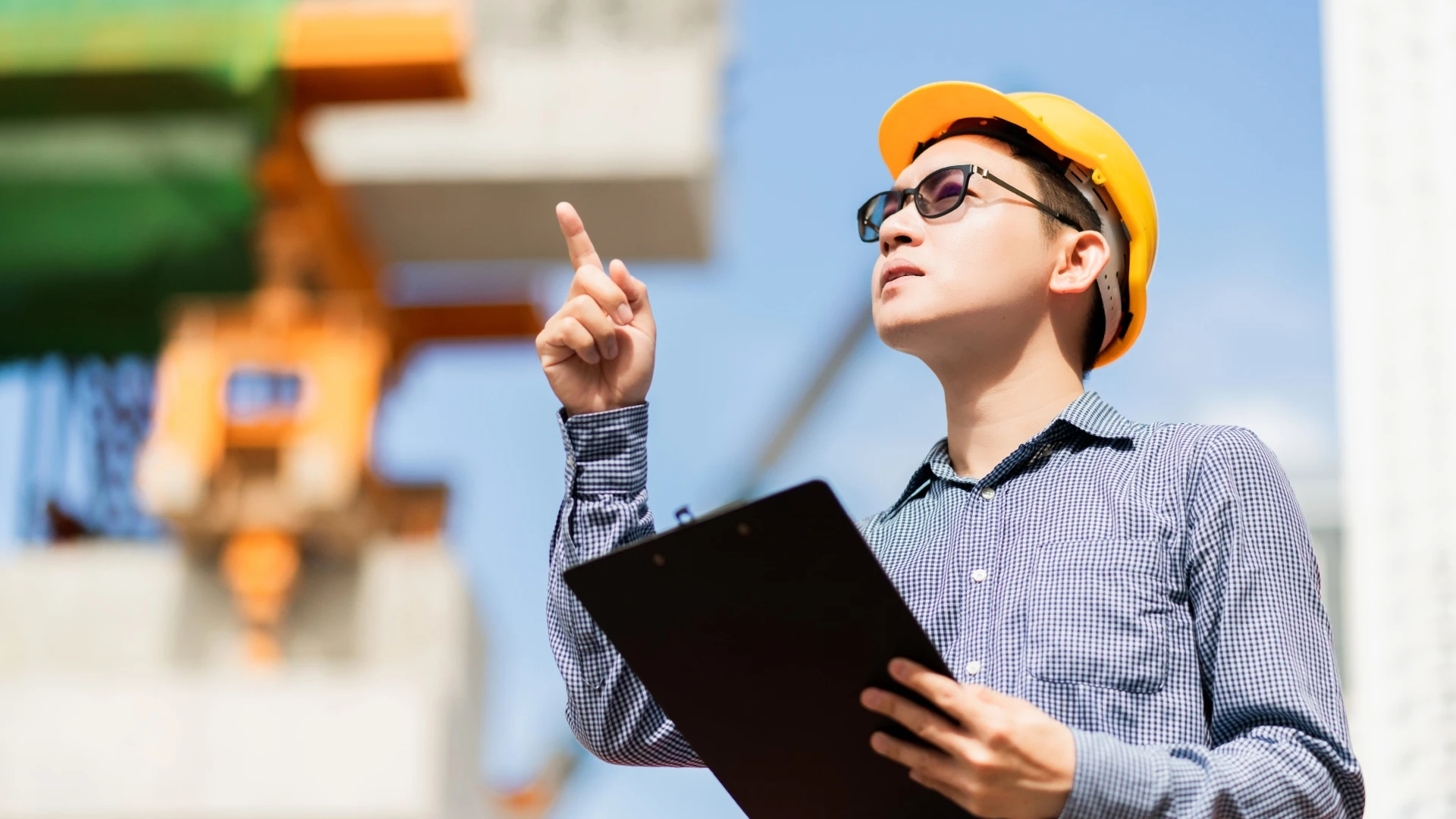 A man wearing a hard hat and holding a clip board points up at a building under construction
