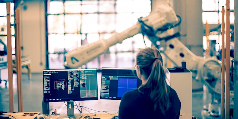 Person observing robotic arm while looking at data on computer.