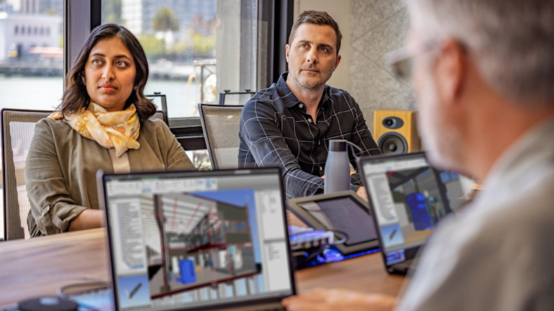 Several people sit around a conference table with open laptops showing architectural plans for a building.
