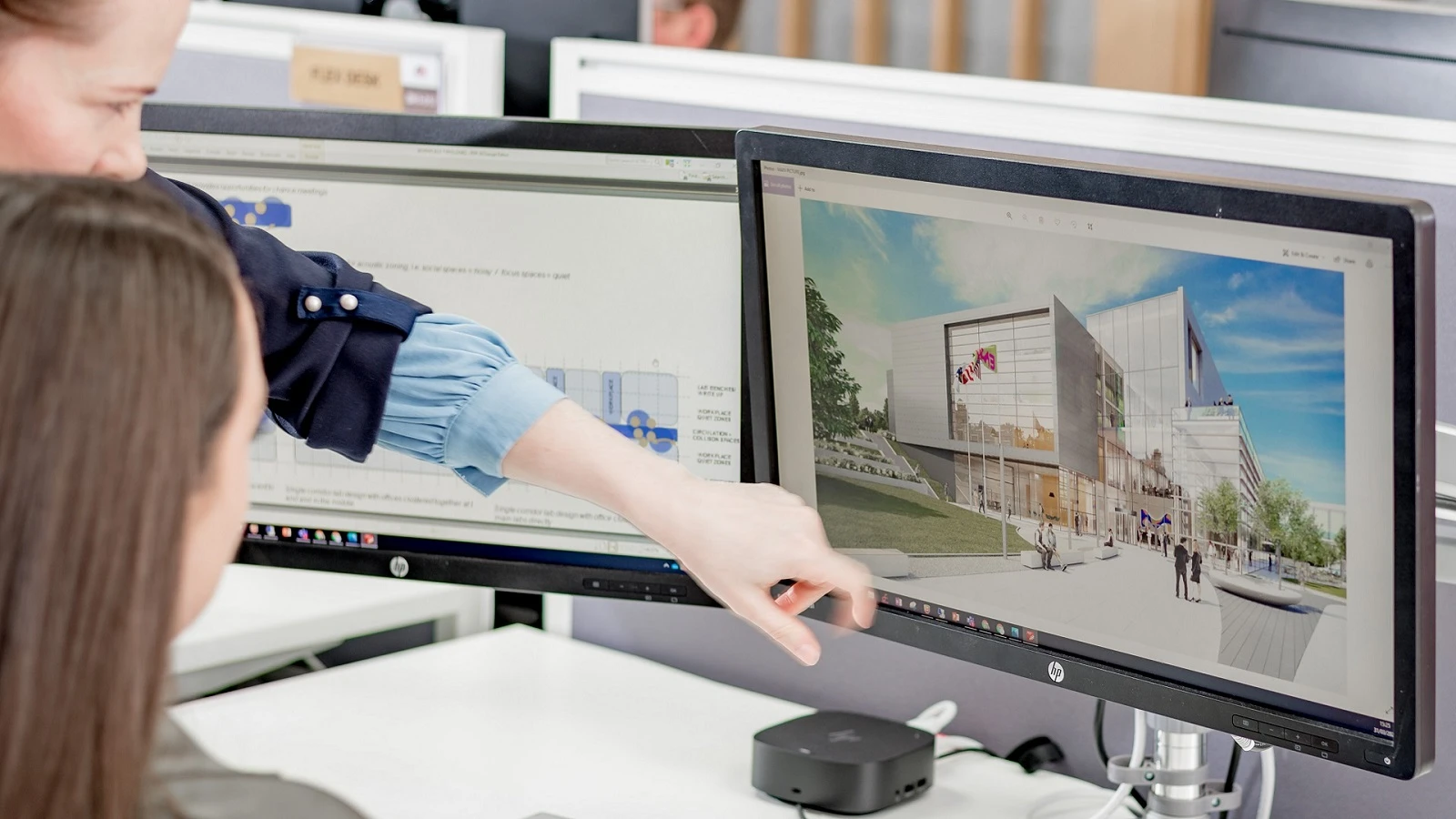 Office workers review a building design on a dual-monitor setup.