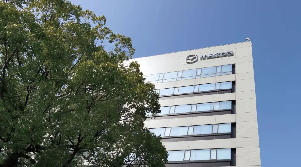 A modern building with the Mazda logo on top, surrounded by greenery under a clear blue sky.
