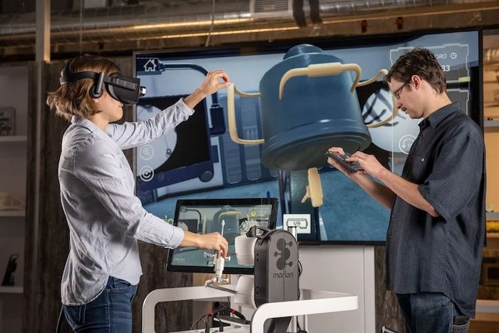 A woman wearing a VR headset interacts with a VR and haptic feedback surgical training platform while her coworker watches.
