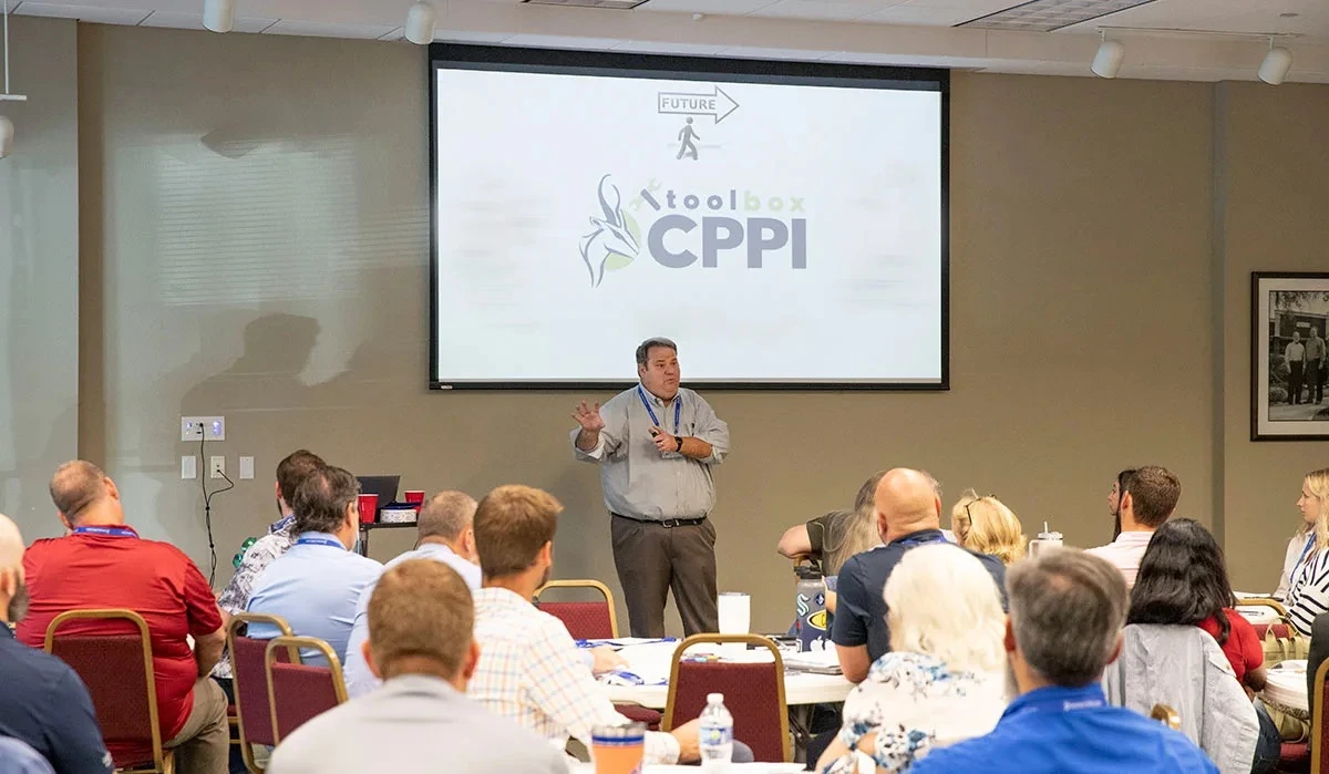 A presenter leads a professional training session as attendees sit at tables facing a projected presentation screen.